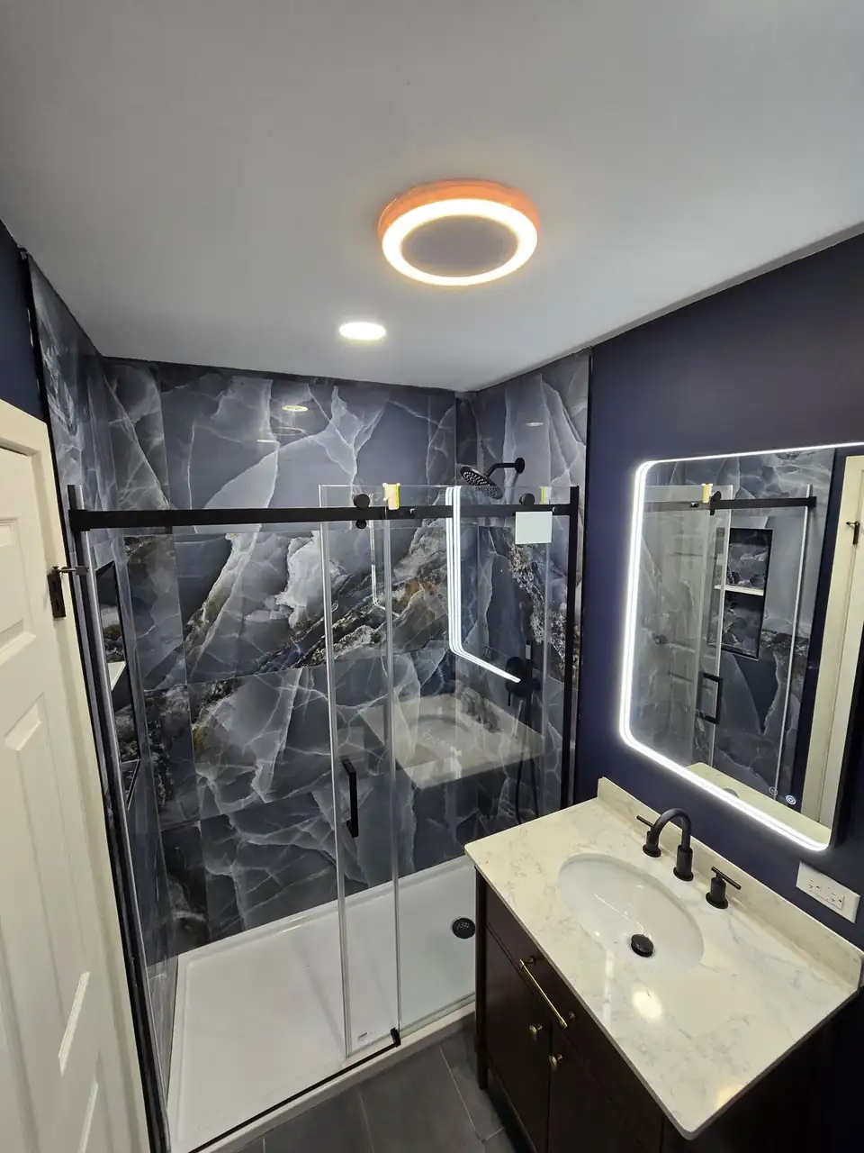 Bathroom with dark marble-look wall panels, a black-framed glass shower door and a lighted mirror: bathroom remodeling in Chicago by Jota Handyman Remodeling