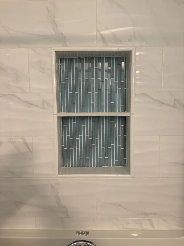 Recessed shower niche lined with green glass mosaic tile in a white marble-look tile wall